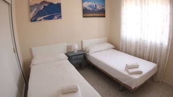 Quarto de hotel com duas camas de solteiro, uma mesa de cabeceira, um abajur fixado na parede e duas imagens emolduradas de montanhas.