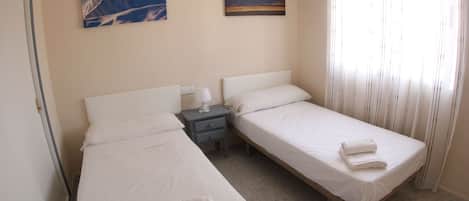 A hotel room with two single beds, a nightstand, a wall-mounted lamp, and two framed pictures of mountains.