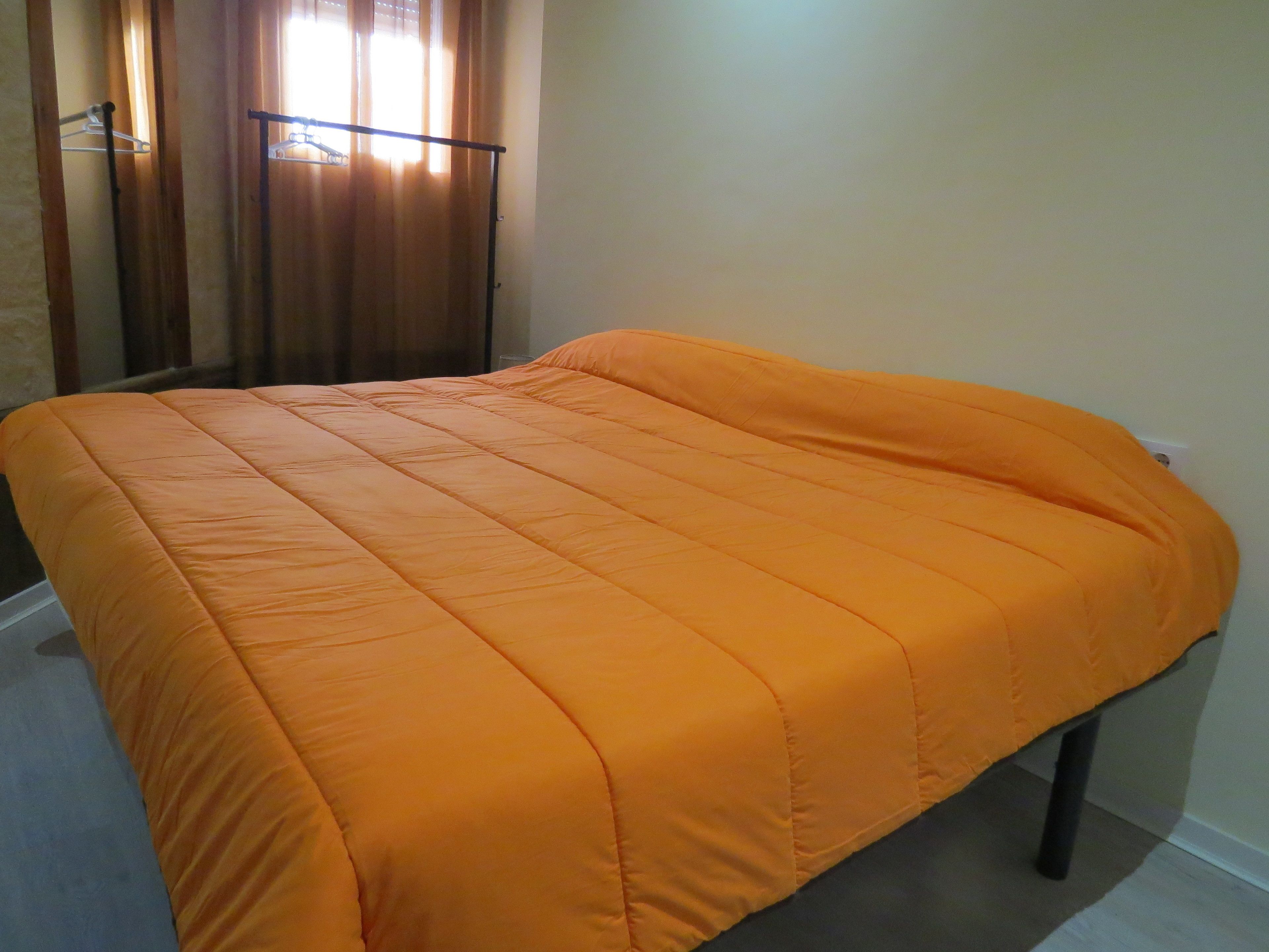 Een bed met een oranje sprei in een kamer met een houten vloer en een raam met gordijnen.
