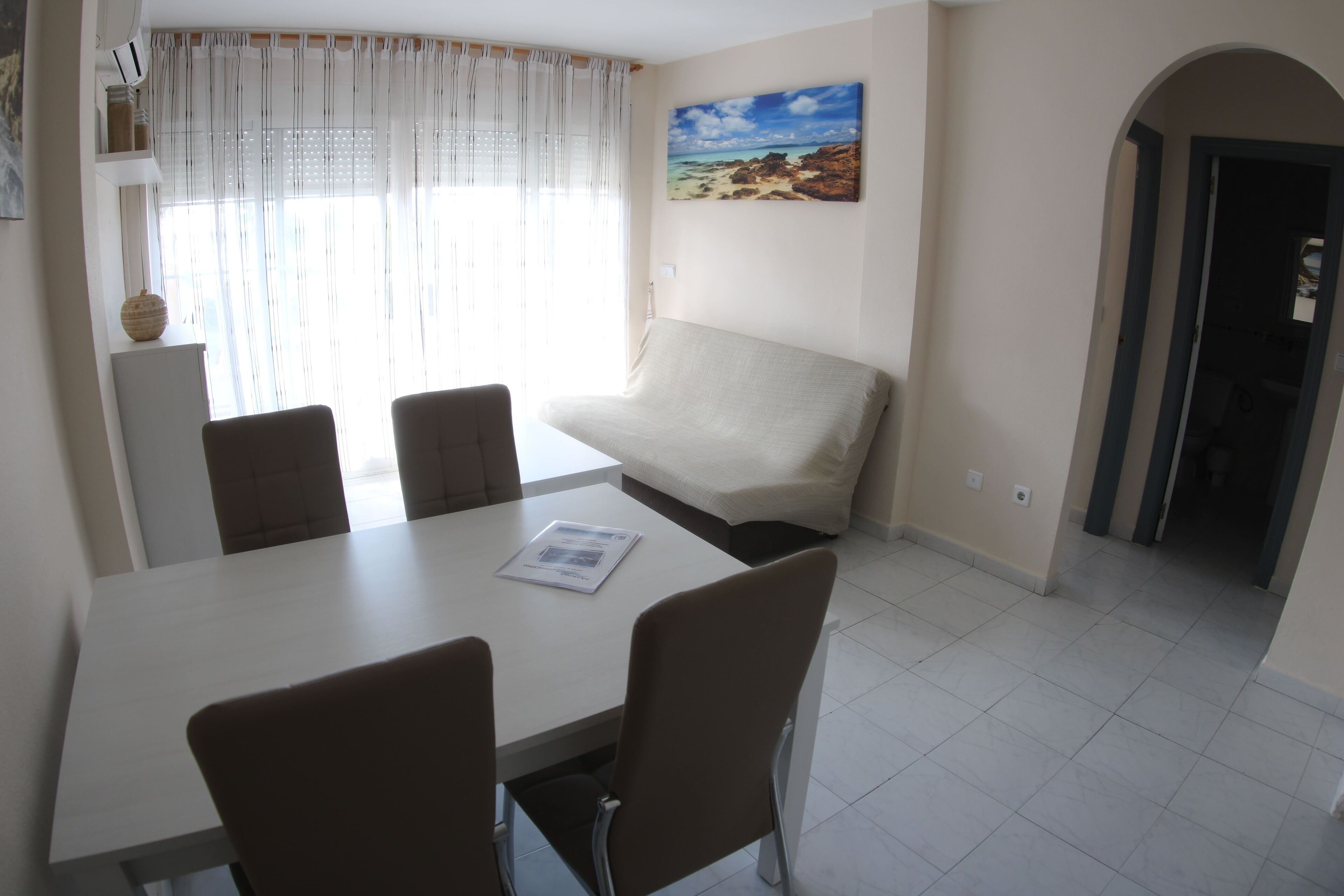 Uma área de refeições com mesa e cadeiras, um sofá, um quadro na parede e vista para a praia pela janela.