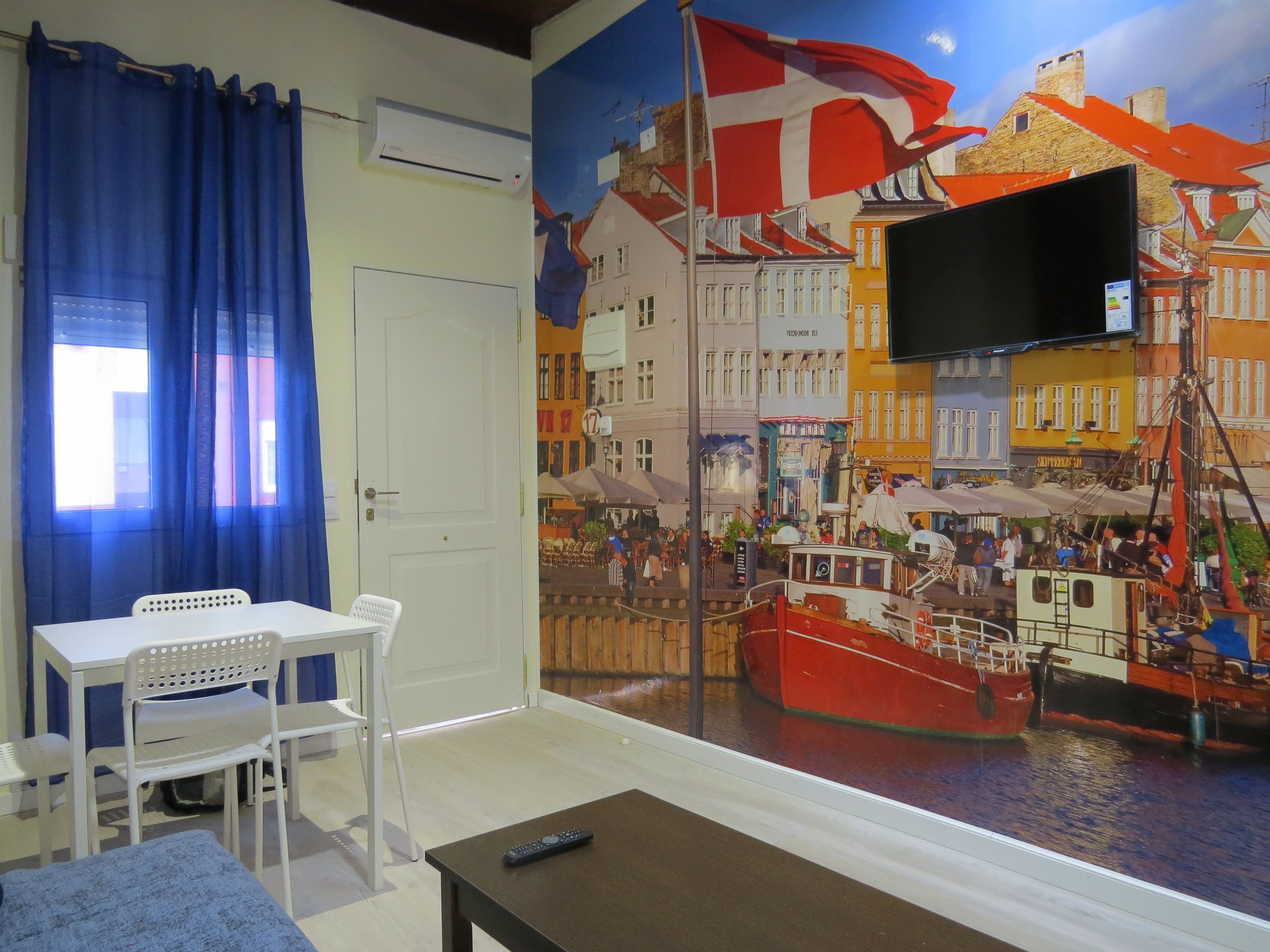 A room with a large window featuring a mural of a bustling harbor scene. There is a white table with two chairs, a small table with a remote control, and a blue curtain.