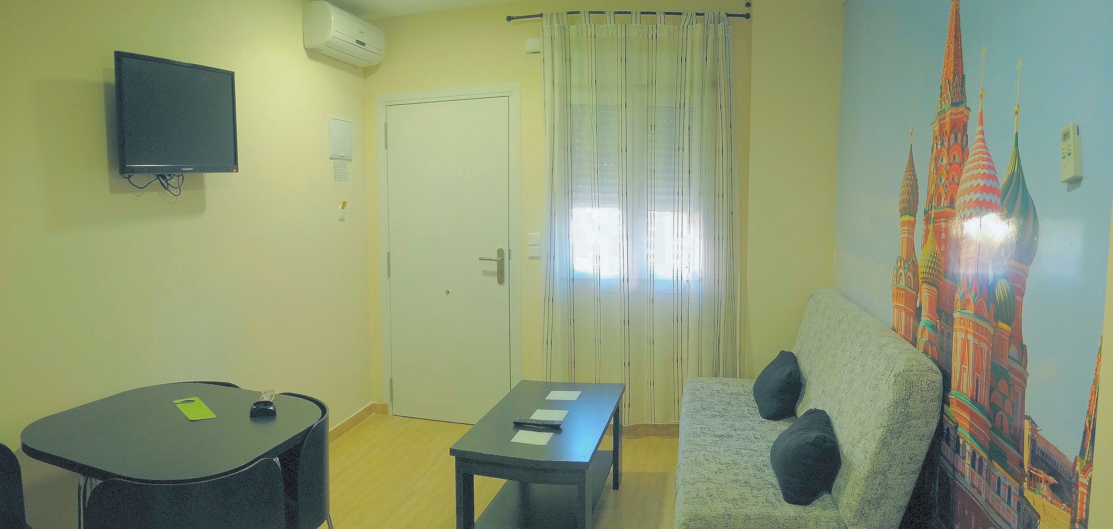 A room with a flat-screen TV mounted on the wall, a door with a curtain, a sofa with cushions, a small table with a chair, and a wall with a colorful mural.