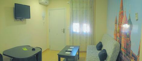 A room with a flat-screen TV mounted on the wall, a door with a curtain, a sofa with cushions, a small table with a chair, and a wall with a colorful mural.