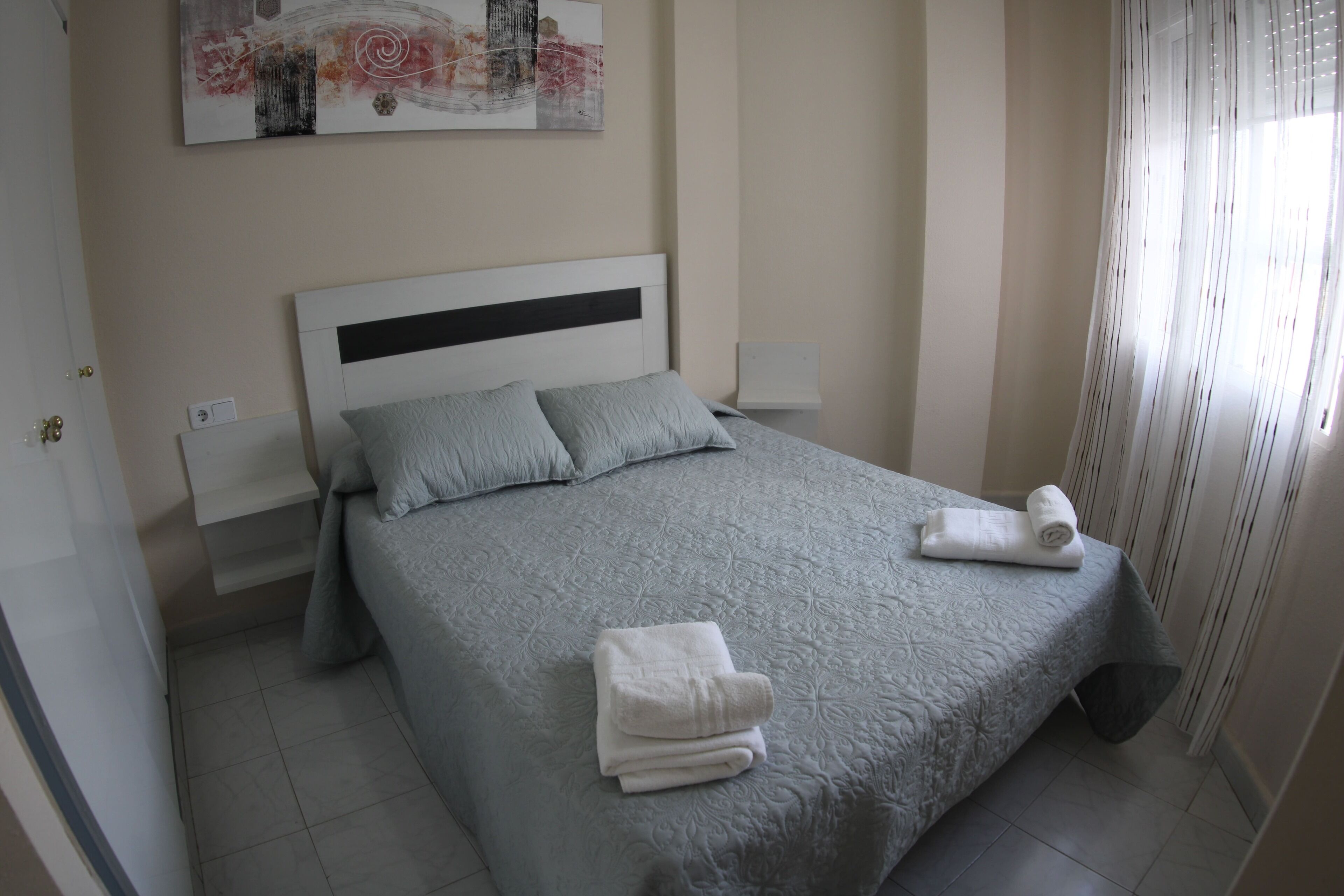 Een netjes opgemaakt bed met een grijze plaid en kussens, een opgevouwen handdoek op het bed, en een schilderij aan de muur erboven.