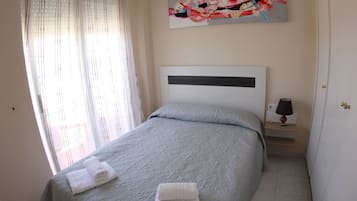 Una cama bien hecha con una colcha gris y dos toallas blancas dobladas, una mesita de noche con una lámpara, y un cuadro abstracto de colores en la pared.