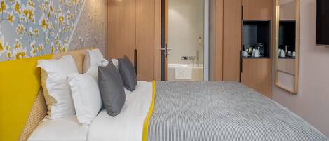 Un dormitorio moderno con una cama, lámparas de noche, un armario y un baño visible a través de una puerta abierta.