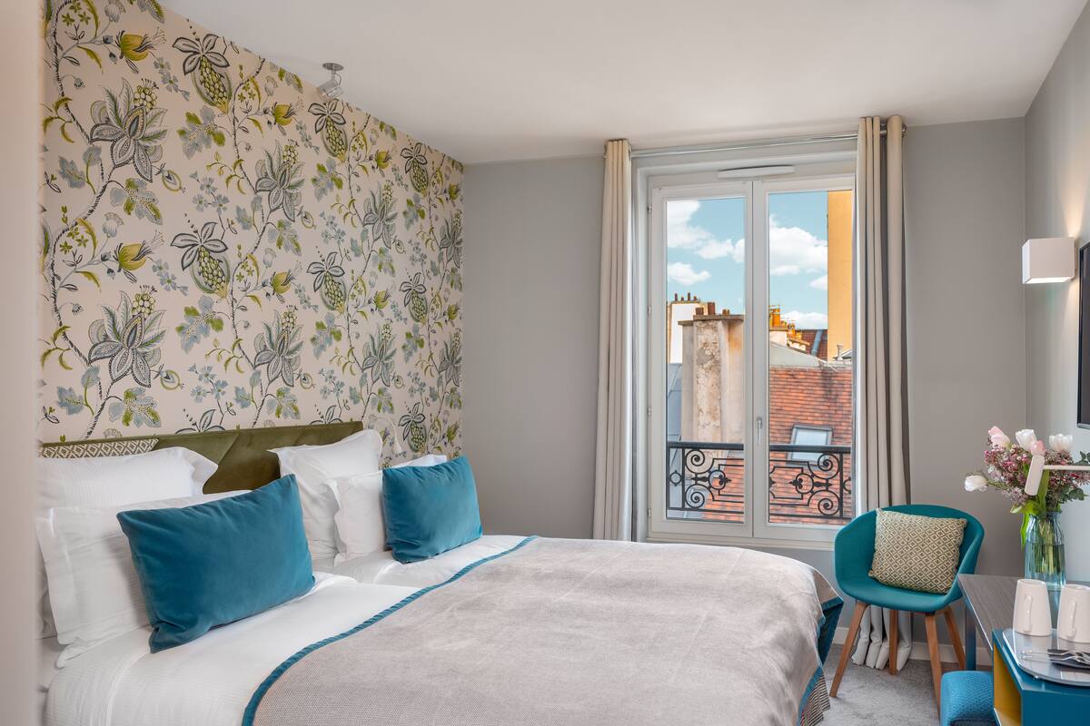 A hotel room with a bed, a chair, a table, and a view of the city through a window.