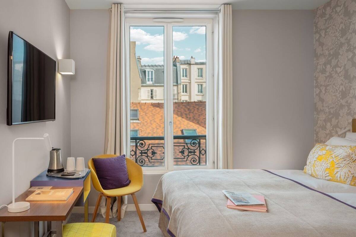 A hotel room with a bed, a desk with a chair, a television, and a window with a view of buildings.