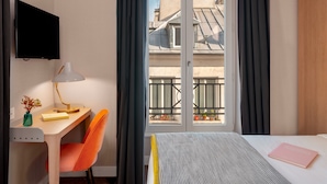 Ett hotellrum med ett skrivbord, en stol, en säng och utsikt över byggnader genom fönstret.