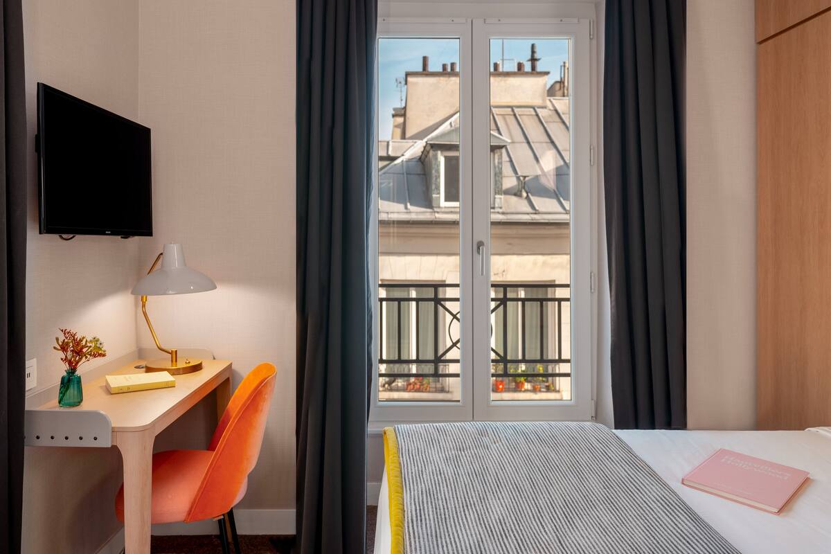 A hotel room with a desk, chair, bed, and a view of buildings through the window.