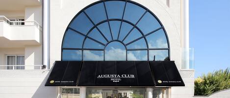 El exterior del Hotel Augusta Club, con un gran ventanal arqueado, un toldo negro y plantas en macetas en la entrada.