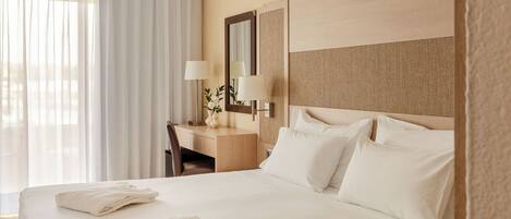 Una habitación de hotel con cama, escritorio, silla, lámpara y ventana con cortinas.