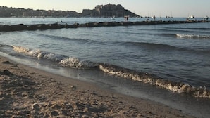 A beach with waves, a distant building, and a clear sky. - Grand Hotel (Calvi)
