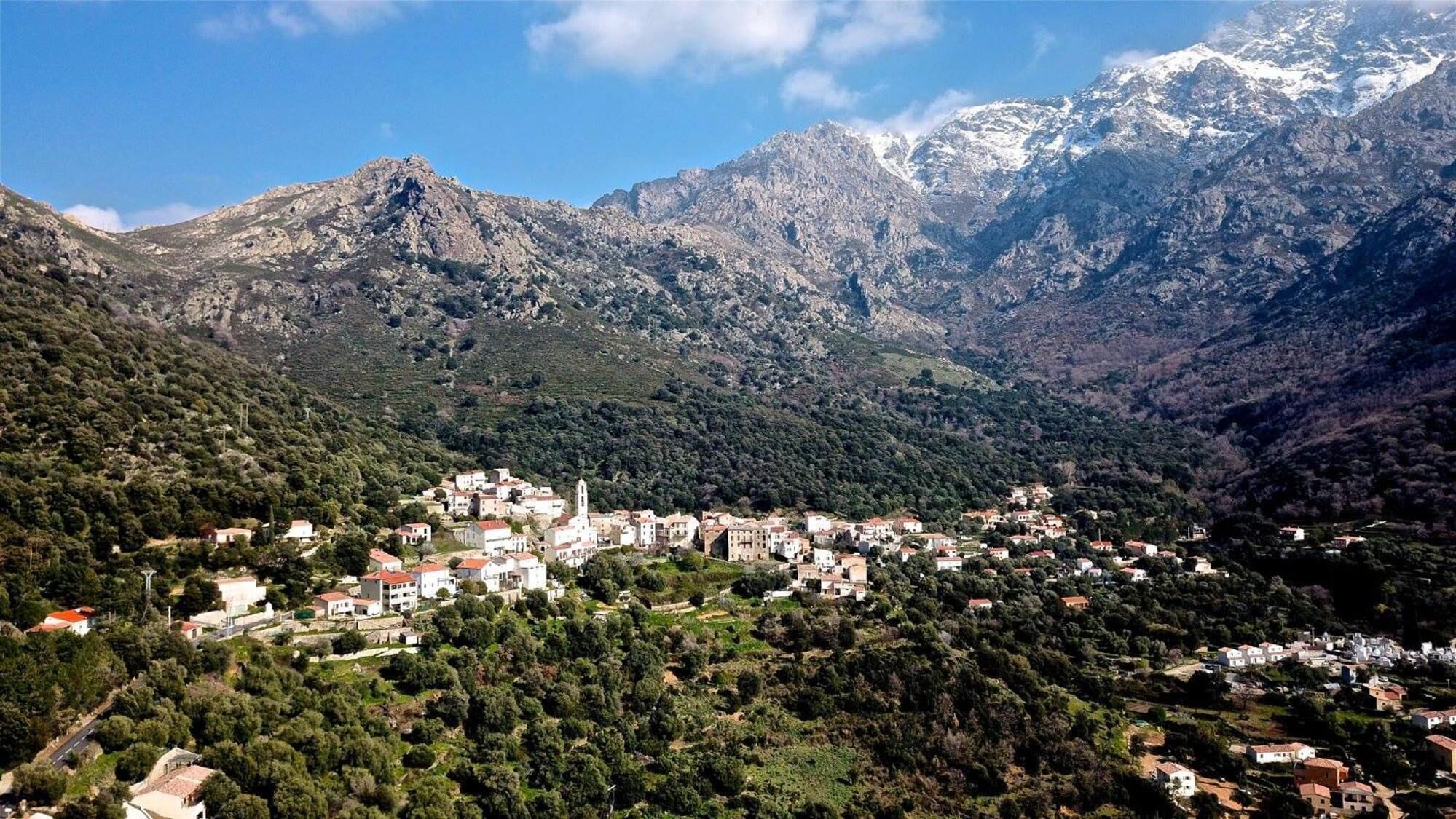 A mountainous village with buildings nestled among the hills, a prominent church, and snow-capped peaks in the background.