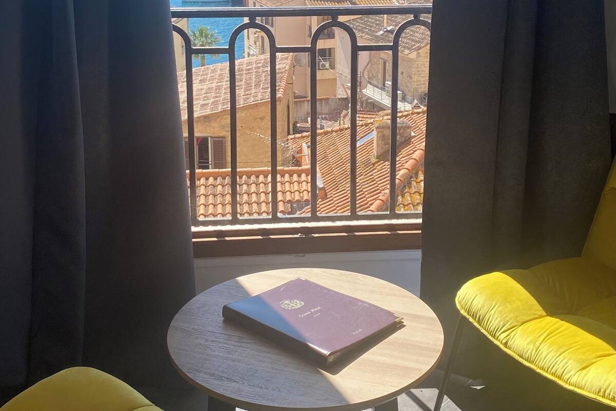 Un balcon avec vue sur un port de plaisance et sur des montagnes. On y trouve une table ronde avec un livre et deux chaises jaunes.
