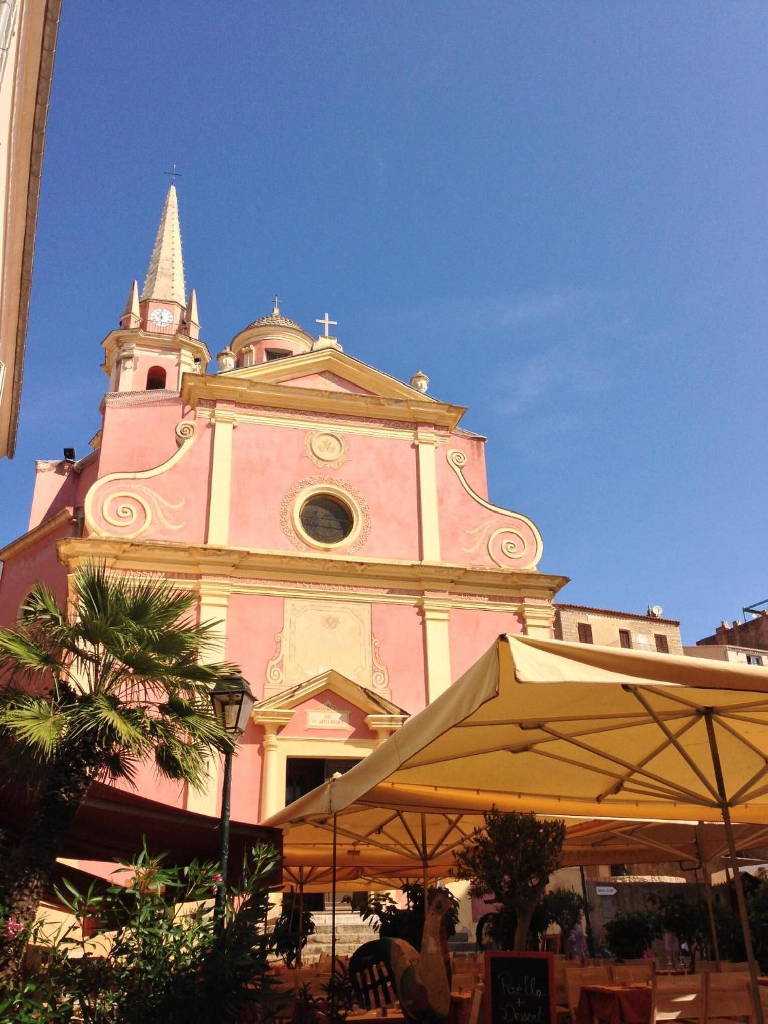 Eine rosa Kirche mit einem Glockenturm und einer Kuppel, umgeben von Außenplätzen und Palmen.