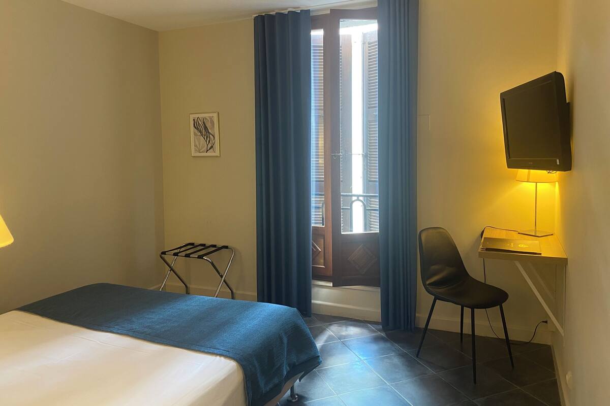 Une chambre d’hôtel comprenant un lit, un bureau avec une chaise, une télévision, une fenêtre avec des rideaux et un tableau accroché au mur.
