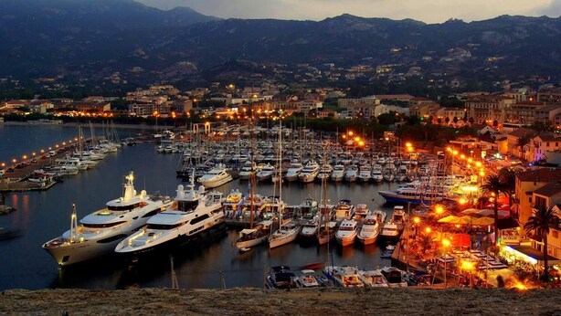 A marina with numerous boats docked, surrounded by buildings and mountains in the background. - Grand Hotel (Calvi)
