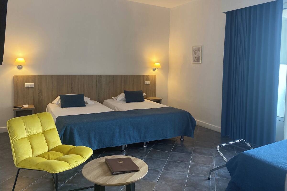 Une chambre d’hôtel avec deux lits, une chaise jaune, une petite table ronde et un rideau bleu.