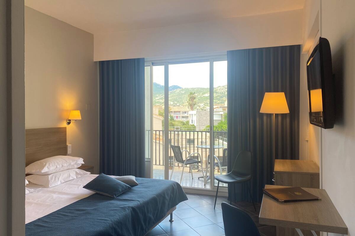 Une chambre d’hôtel avec un lit, un bureau, un ordinateur portable et un balcon équipé de chaises.