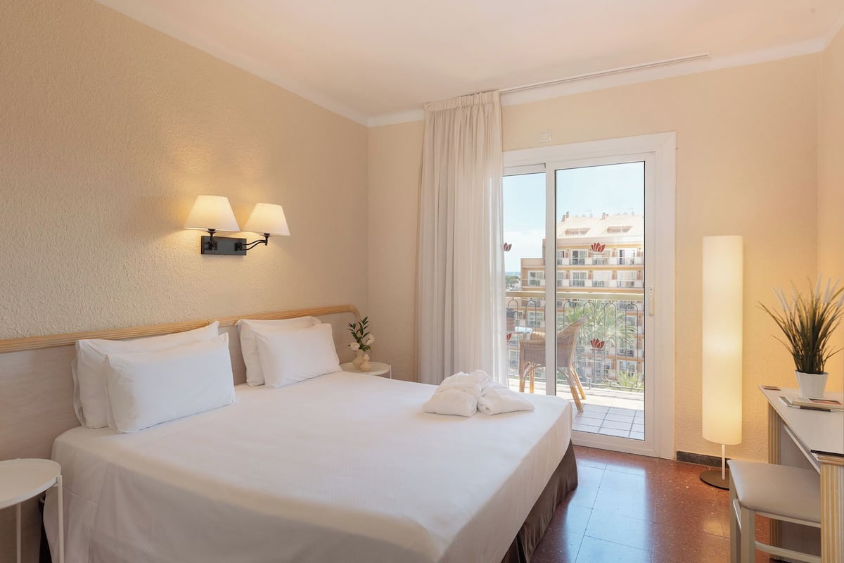 Une chambre d’hôtel avec un lit, deux lampes fixées au mur, un bureau avec une lampe, une plante en pot et une vue sur les bâtiments à travers une porte coulissante.