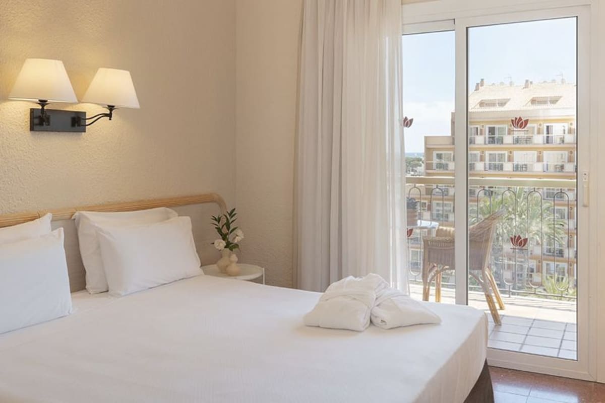 Quarto de hotel com cama, abajures, janela com vista para edifícios e uma planta em vaso sobre uma mesa.