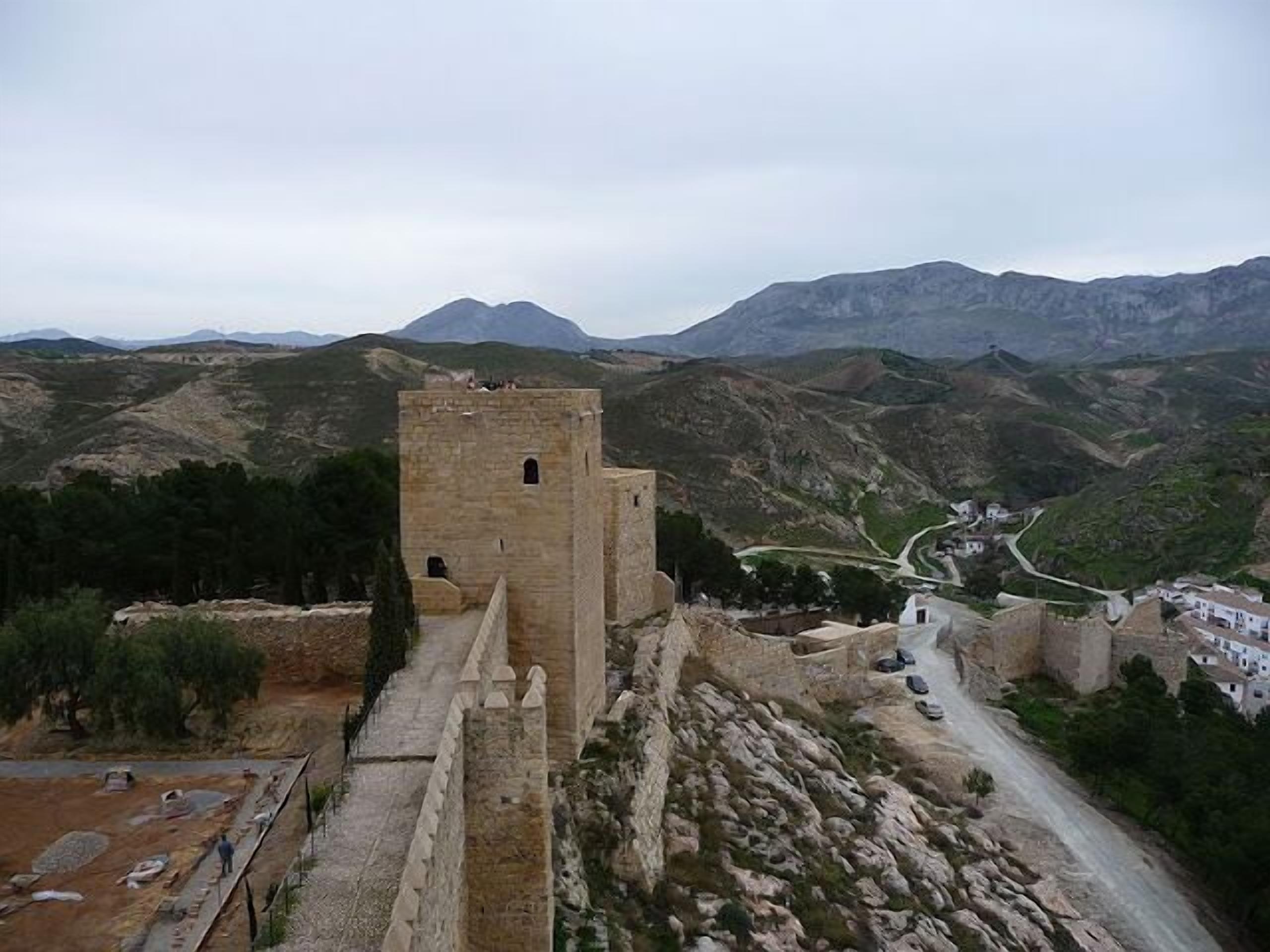 Una torre de piedra histórica con un sendero estrecho que la lleva hasta ella, rodeada de terreno accidentado y montañas a la distancia.