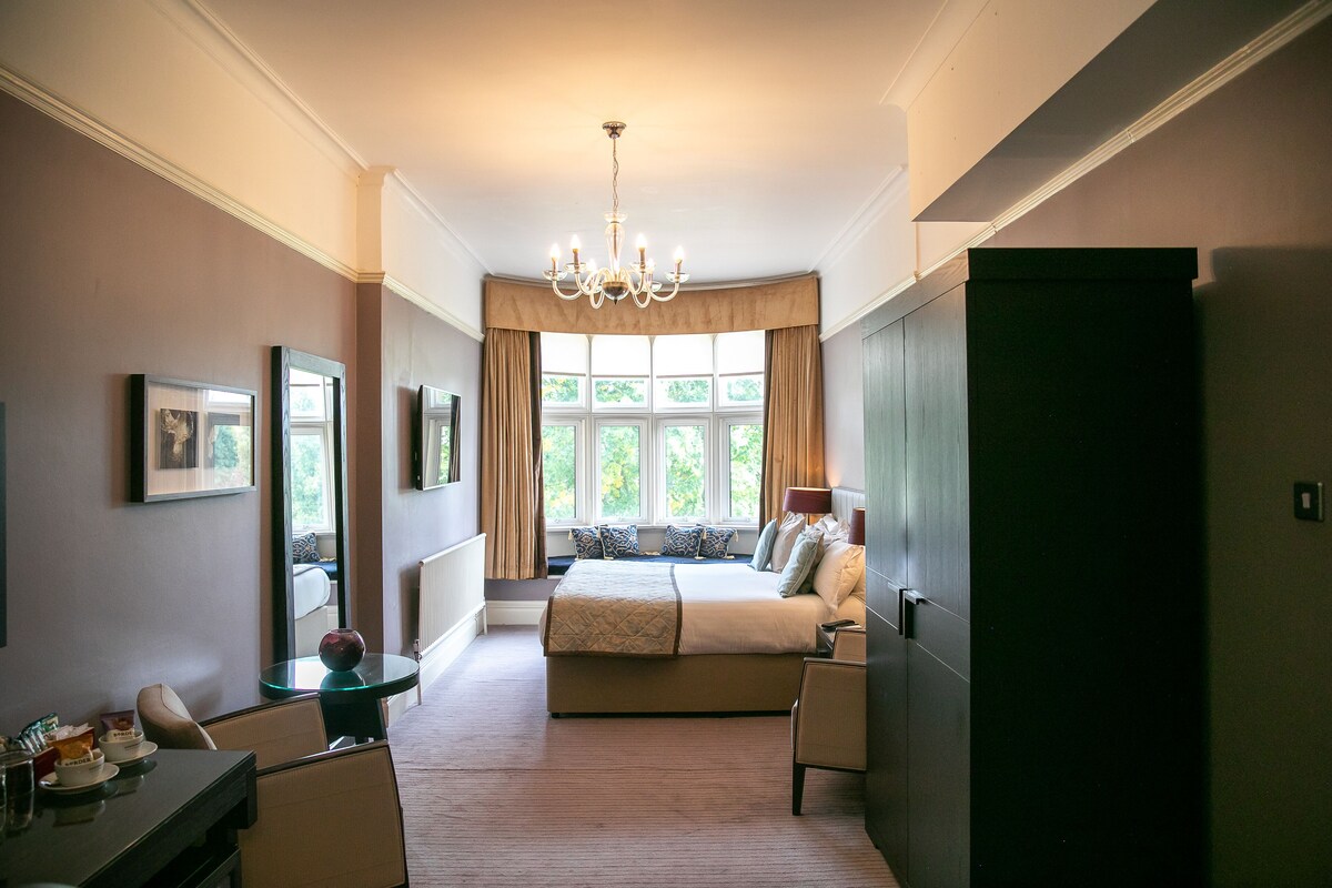 A bedroom with a large bed, a sitting area with a table and chairs, a chandelier, and a view of greenery through the windows.