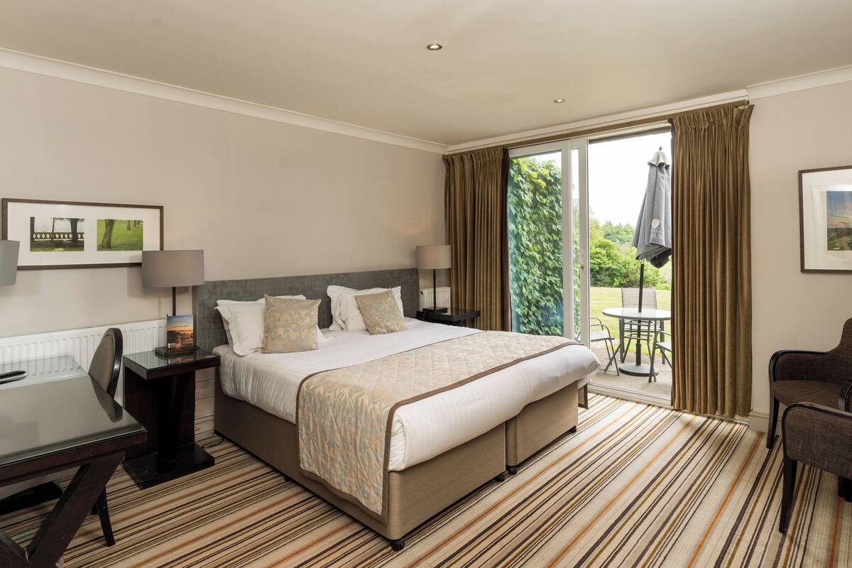 A hotel room with a large bed, a desk with a chair, a small table, and a view of the outdoors through a sliding door.