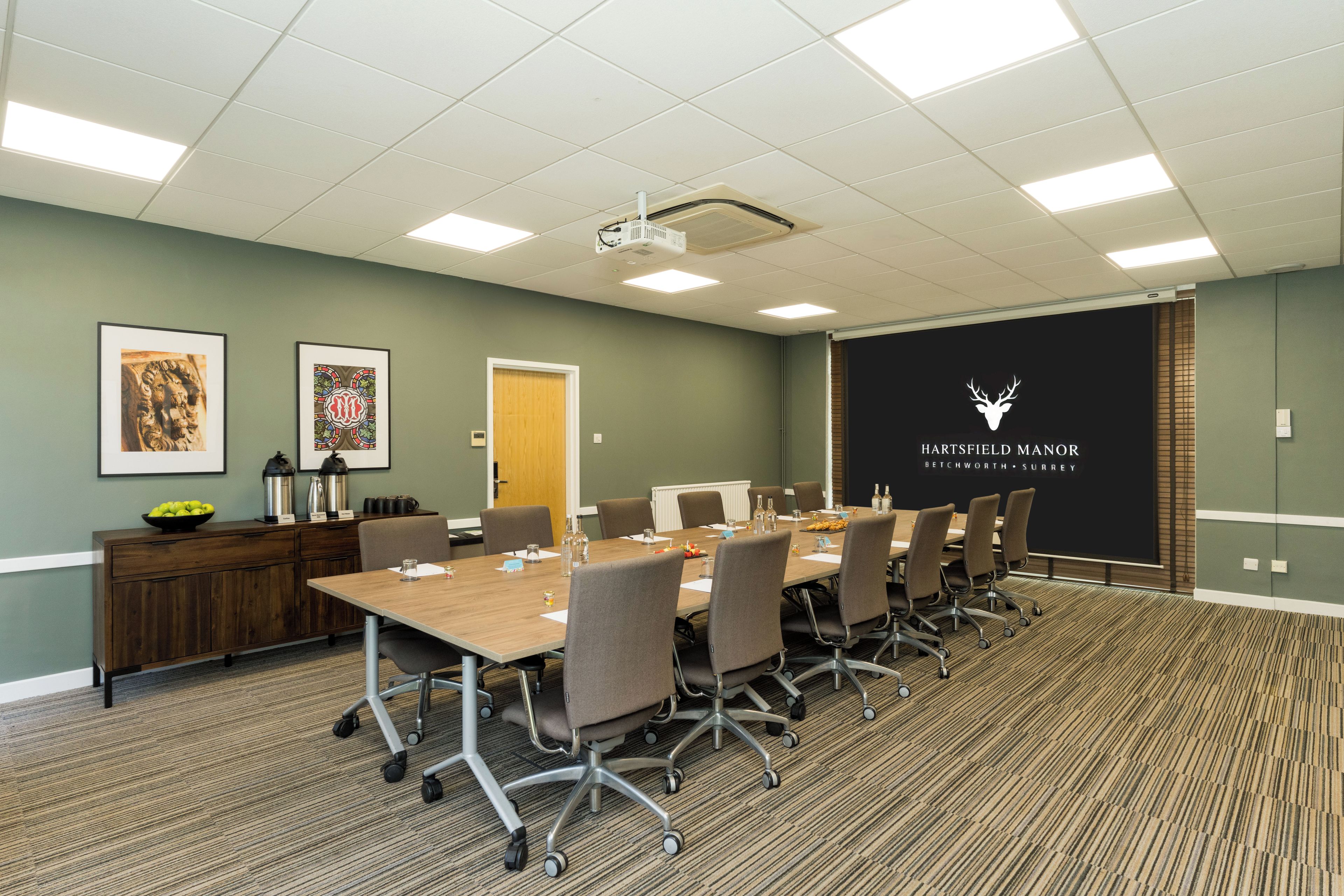 A conference room with a long table, chairs, a projector, and a screen with the logo 'HARTFIELD MANOR BETCHWORTH - SURREY'.