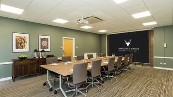 A conference room with a long table, chairs, a projector, and a screen with the logo 'HARTFIELD MANOR BETCHWORTH - SURREY'.