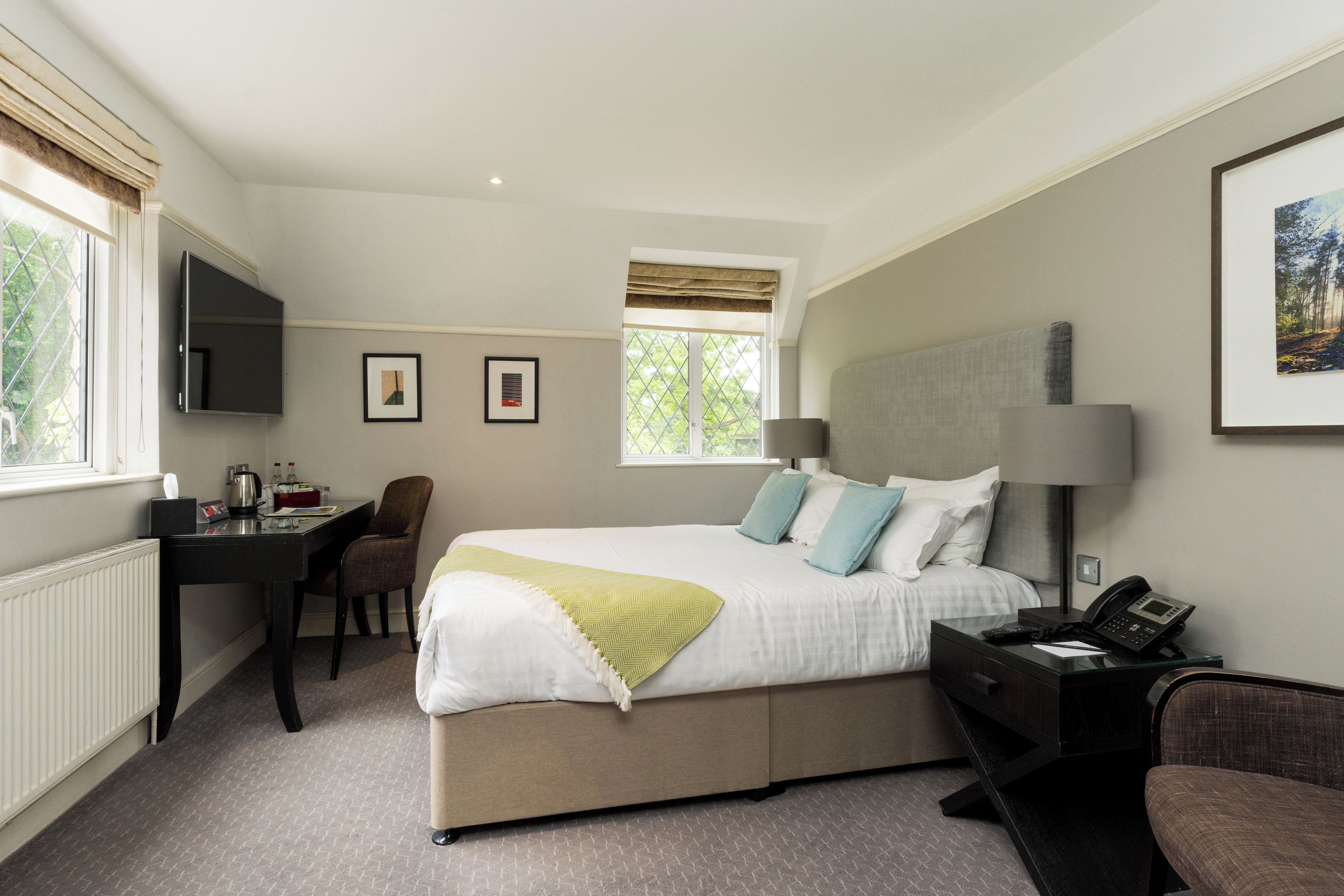 Hotelkamer met een bed, nachtkastje, lamp, stoel, bureau, televisie en ingelijste foto's aan de muur.