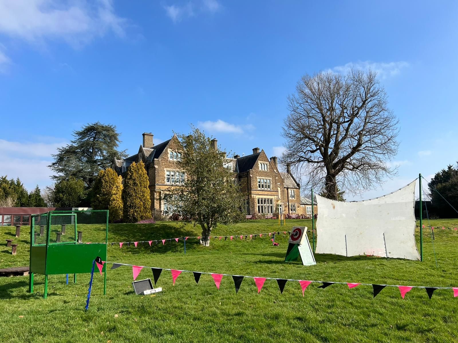 A grassy area with a large house in the background, a green structure, and a white tent-like object.