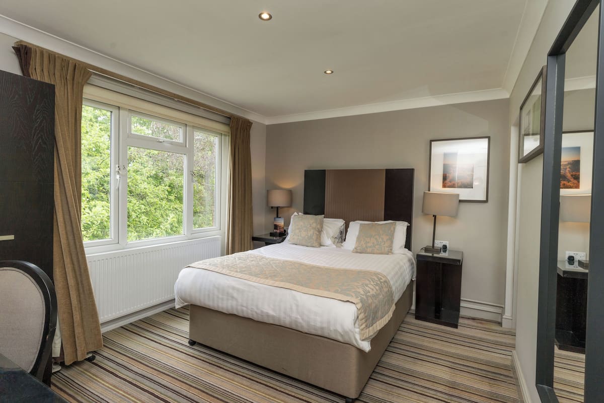 A hotel room with a large bed, two bedside tables with lamps, a window with curtains, and a framed picture on the wall.