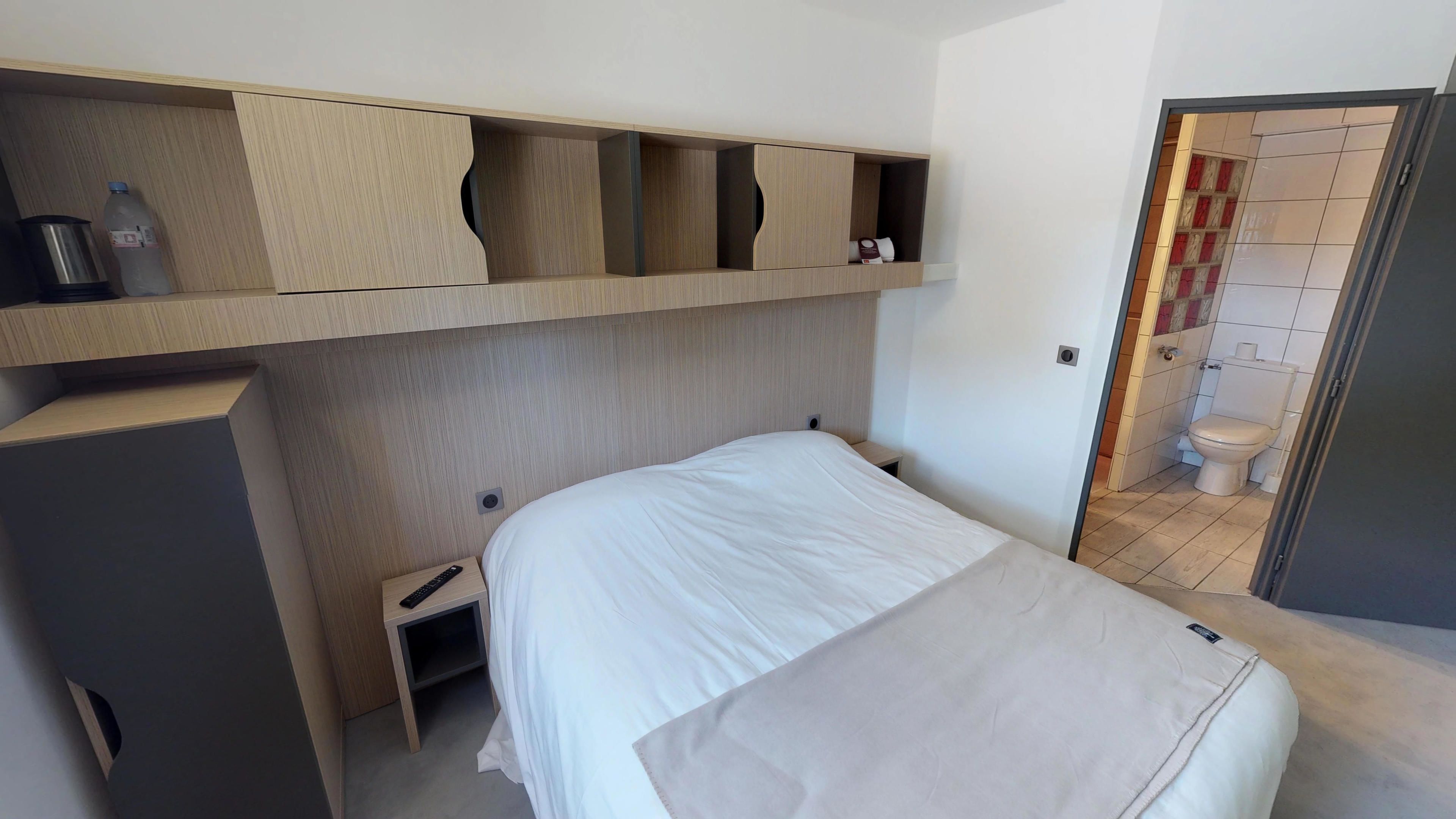 A compact hotel room with a bed, a small bedside table, a built-in shelf with storage compartments, and a view of the bathroom.