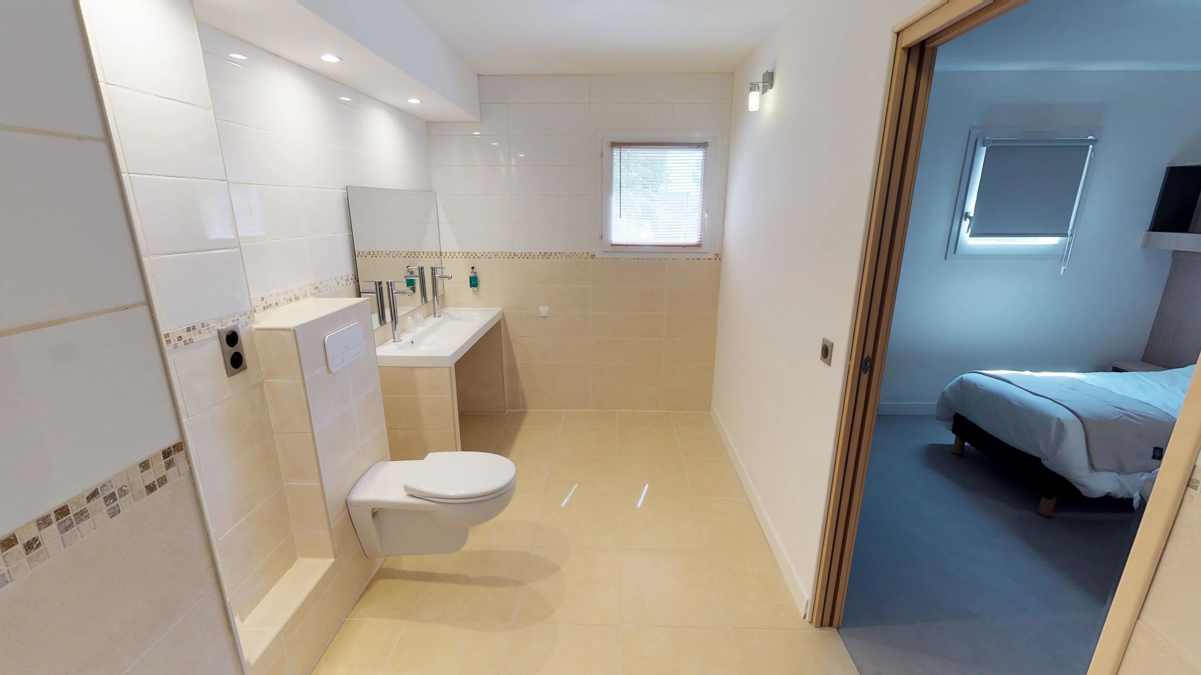 Une salle de bain moderne équipée de toilettes, d’un lavabo et d’une douche. Une fenêtre avec des stores et une porte donnant sur une chambre à coucher avec un lit sont également présentes.