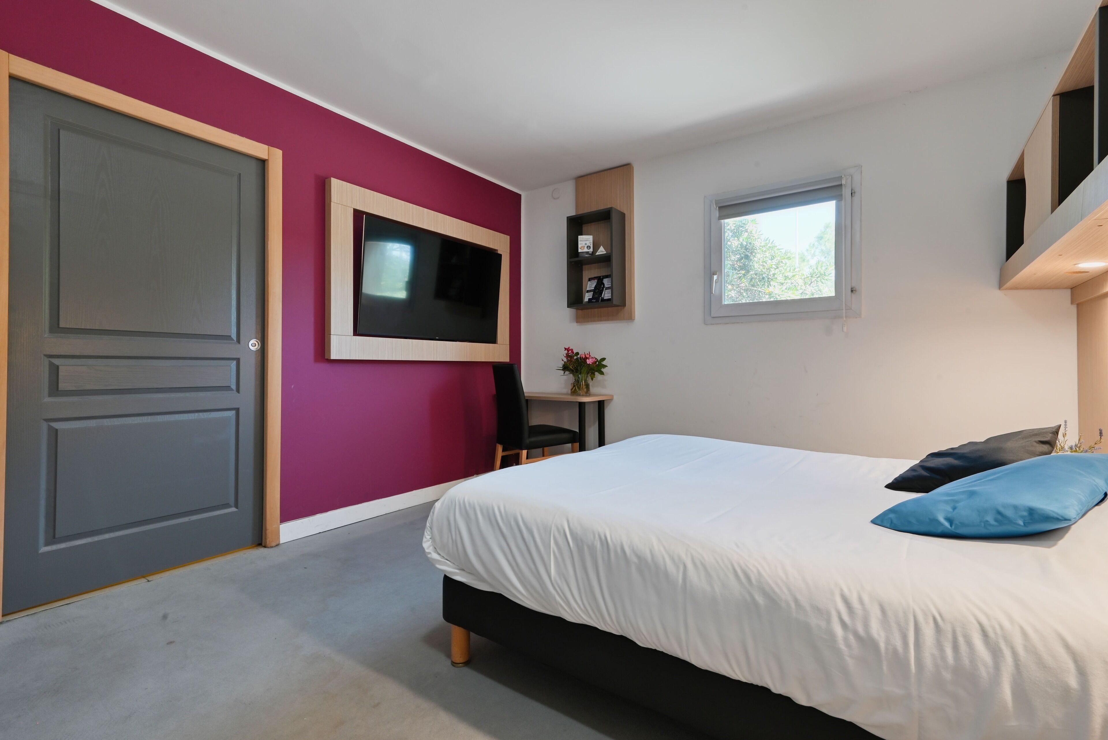 A bedroom with a bed, a TV mounted on the wall, a window, and a door.