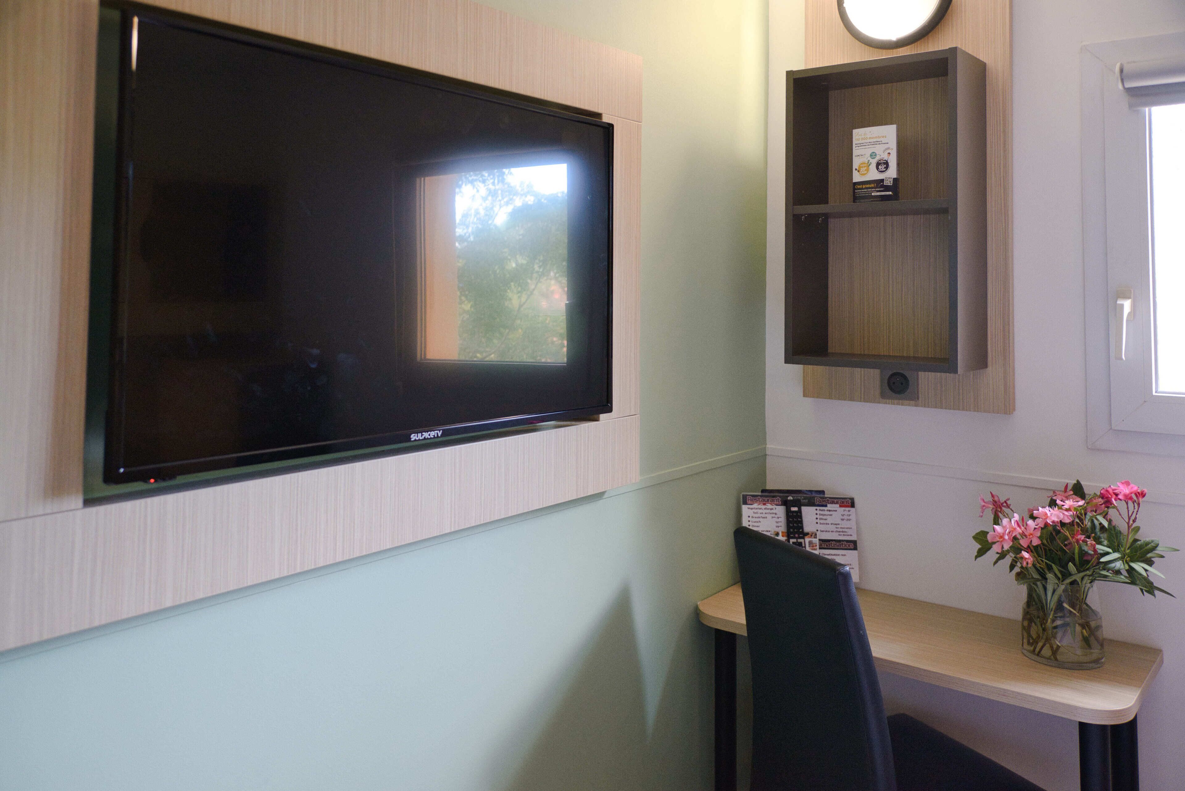 A modern room with a mounted flat-screen TV, a small shelf with a book, a vase with flowers, and a chair.