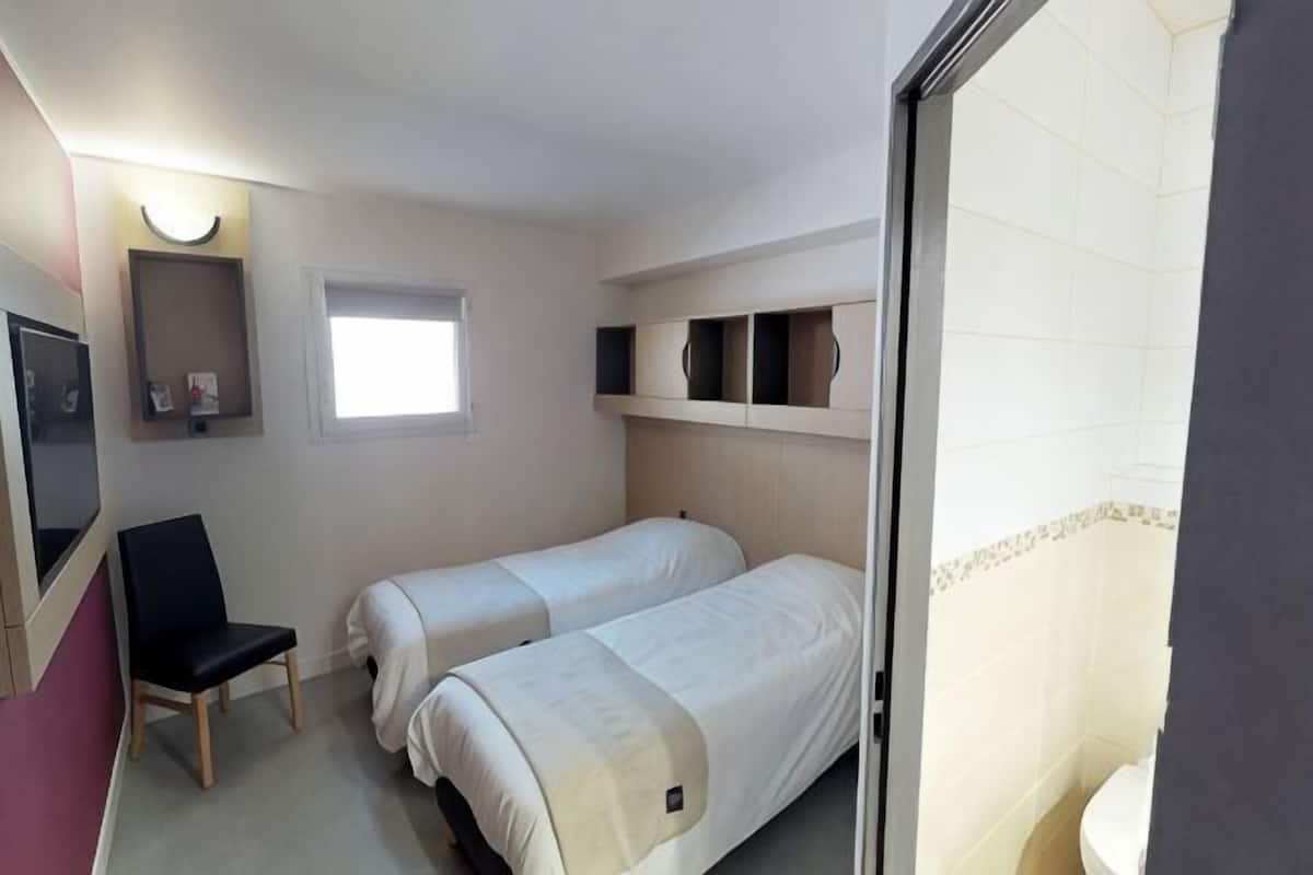 Une chambre avec deux lits, une chaise, une petite fenêtre et une salle de bain visible à travers une porte ouverte.