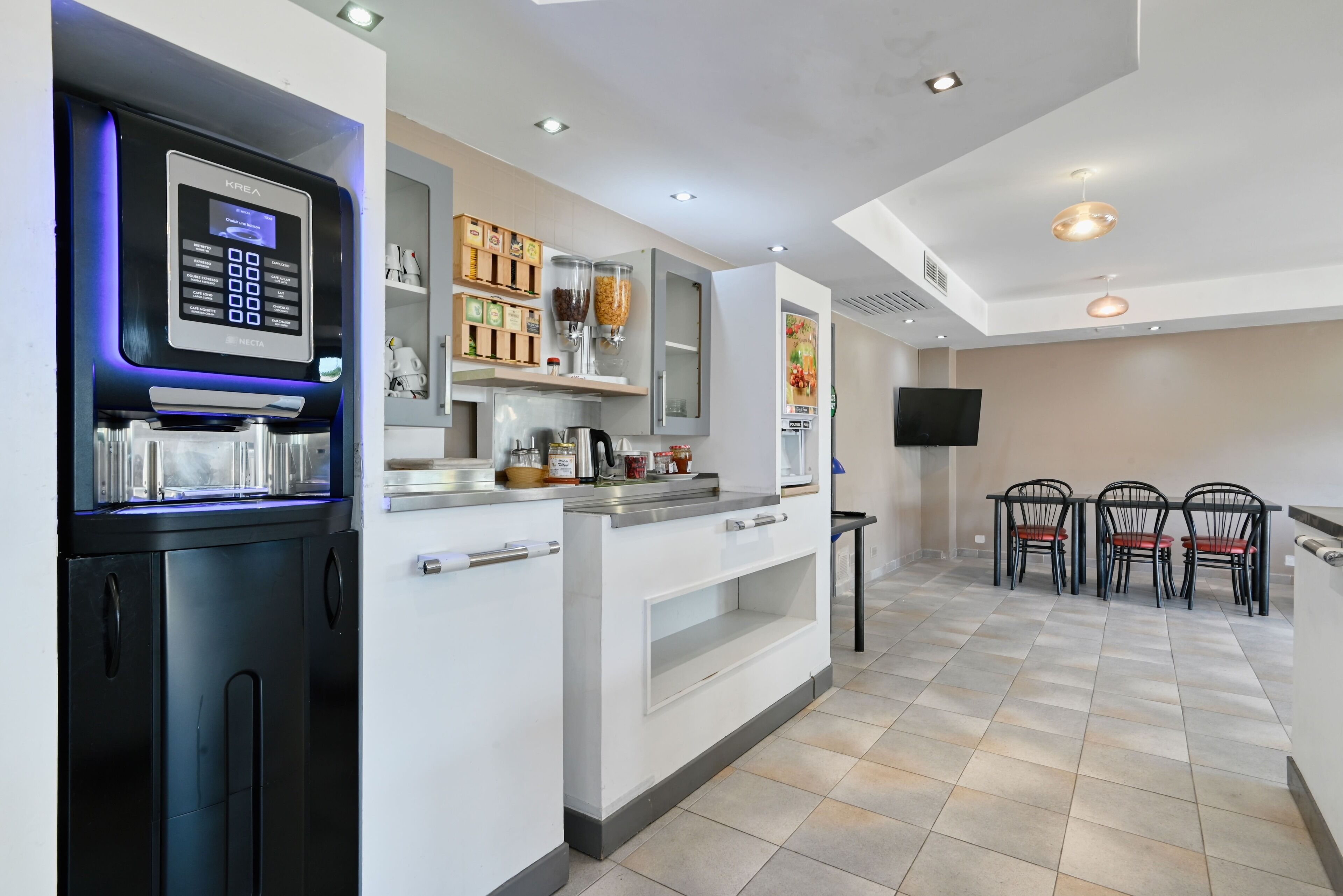 Una cocina moderna con cafetera integrada, microondas y hervidor. Hay armarios con utensilios de cocina y una pequeña zona de comedor con sillas.