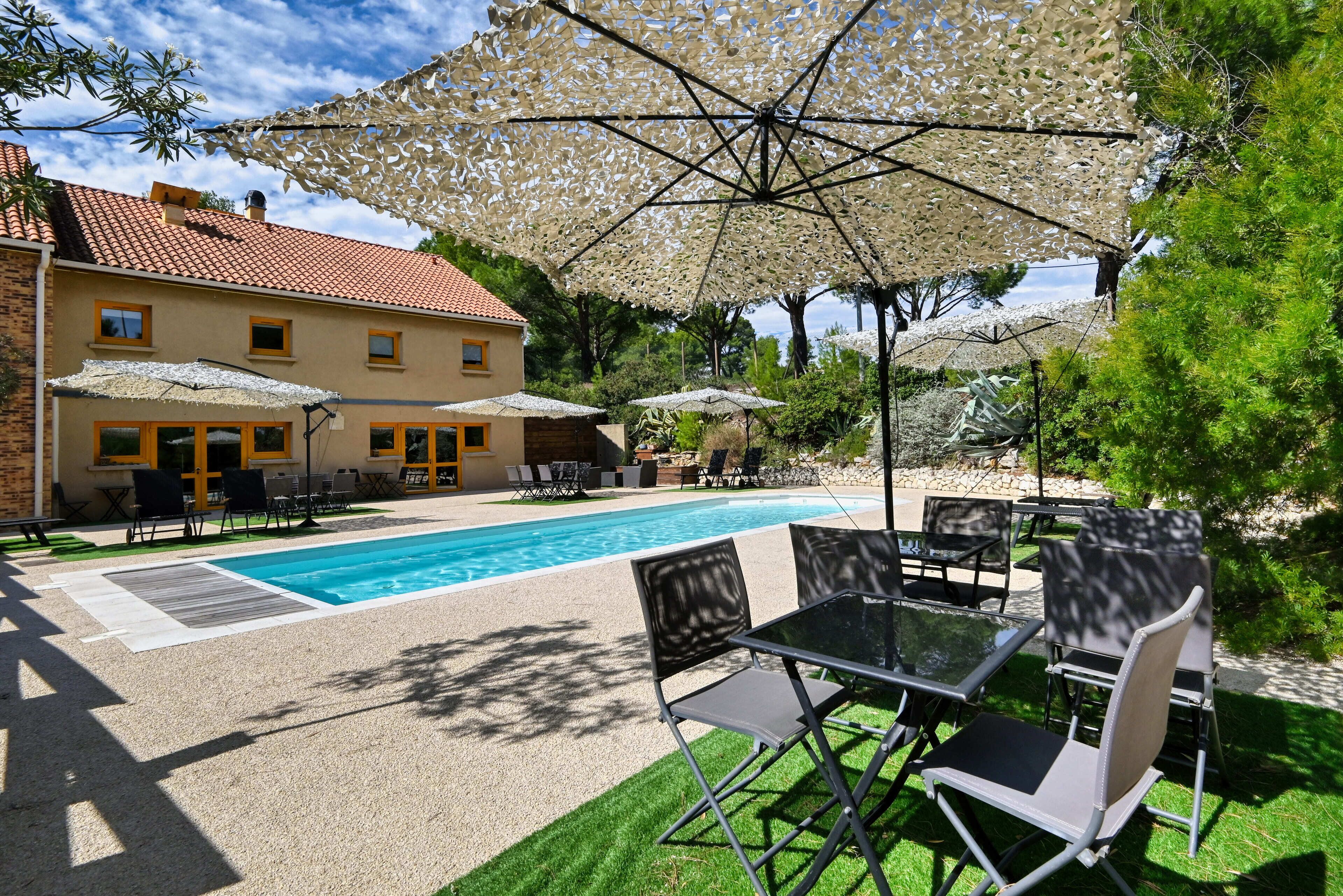 Zona de piscina con mobiliario exterior y sombrillas, piscina y un edificio con tejado de baldosas.
