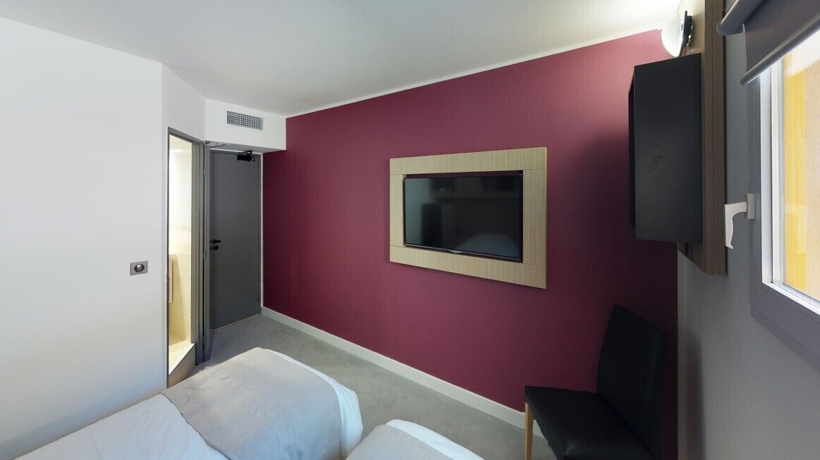 A hotel room with a bed, a chair, a TV mounted on the wall, and a door leading to another room.
