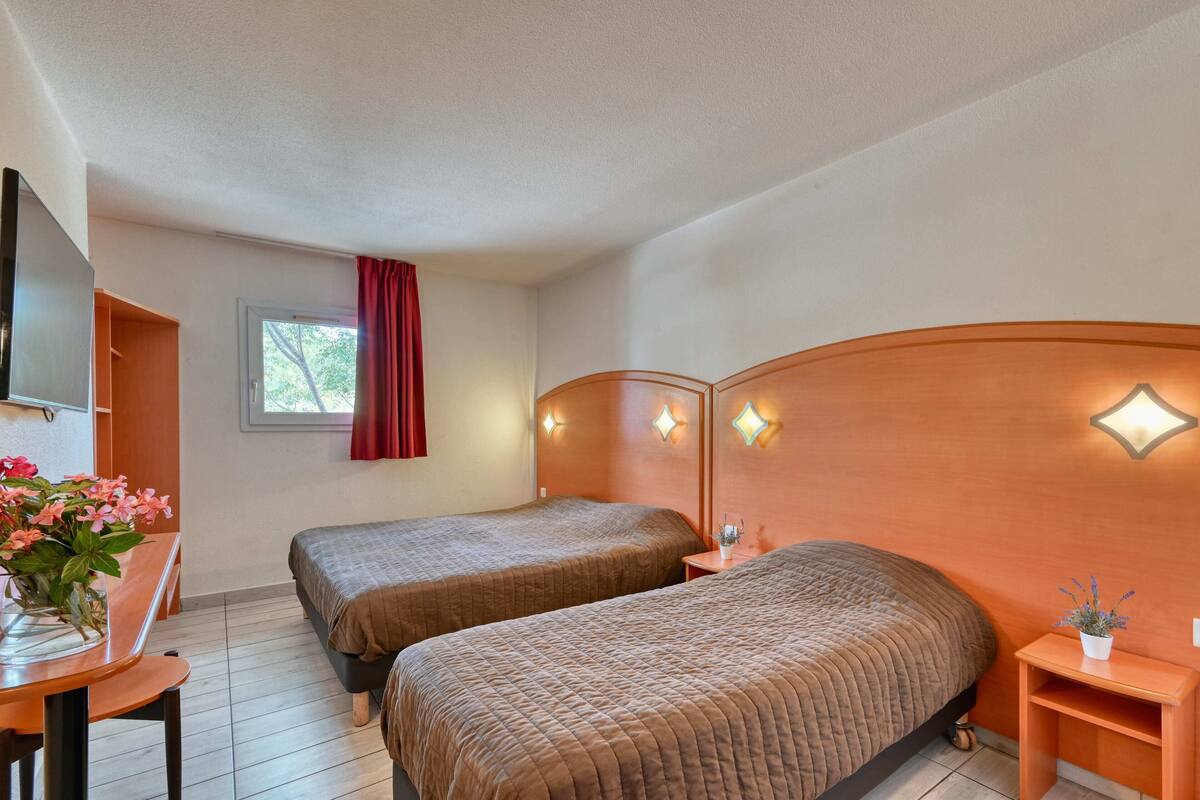 Une chambre avec deux lits, un bureau, une chaise, une fenêtre avec des rideaux rouges et des lampes fixées au mur.