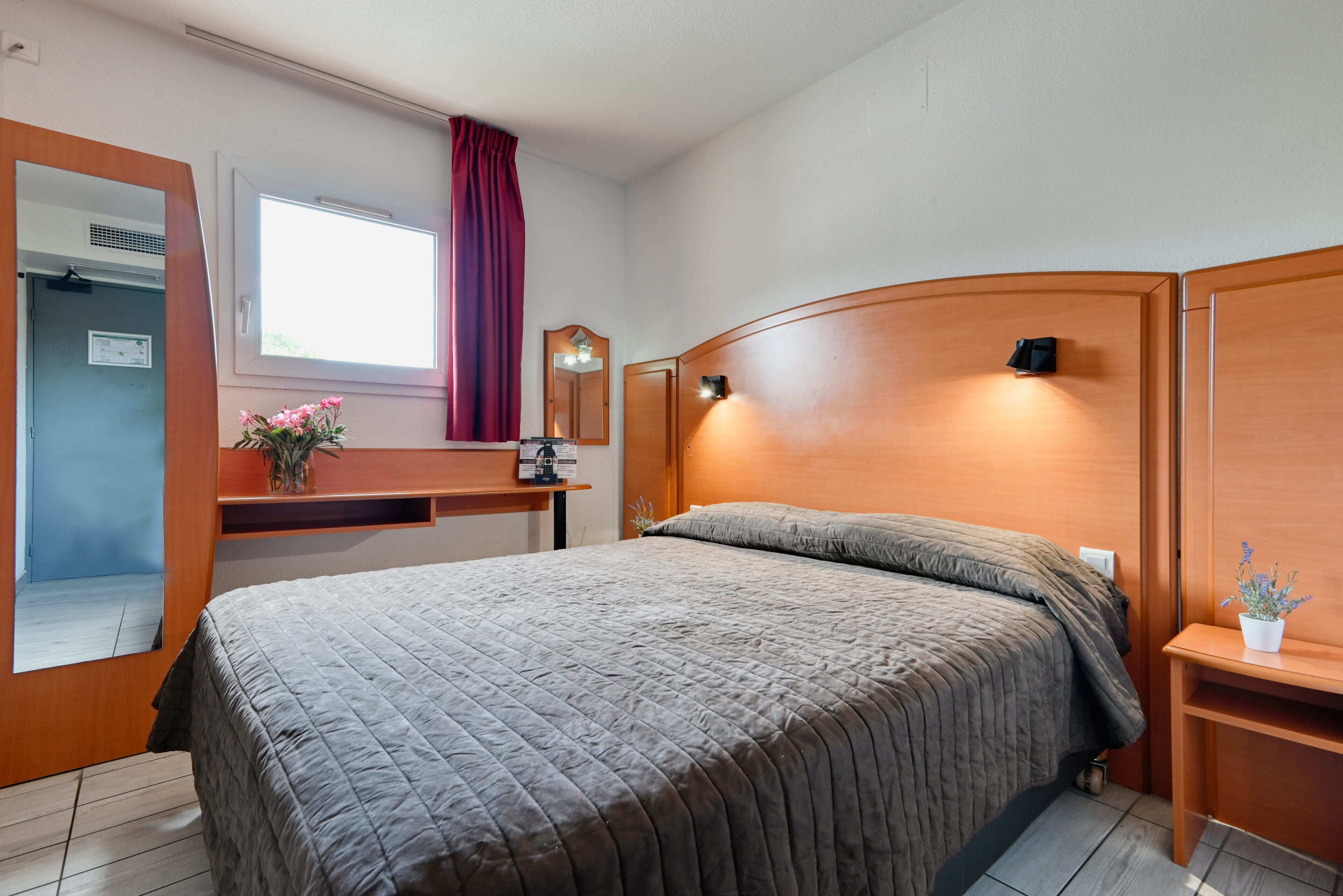 Ein Schlafzimmer mit einem Bett, einem hölzernen Kopfteil, einem Nachttisch mit Pflanze, einem Spiegel und einem Fenster mit Vorhängen.