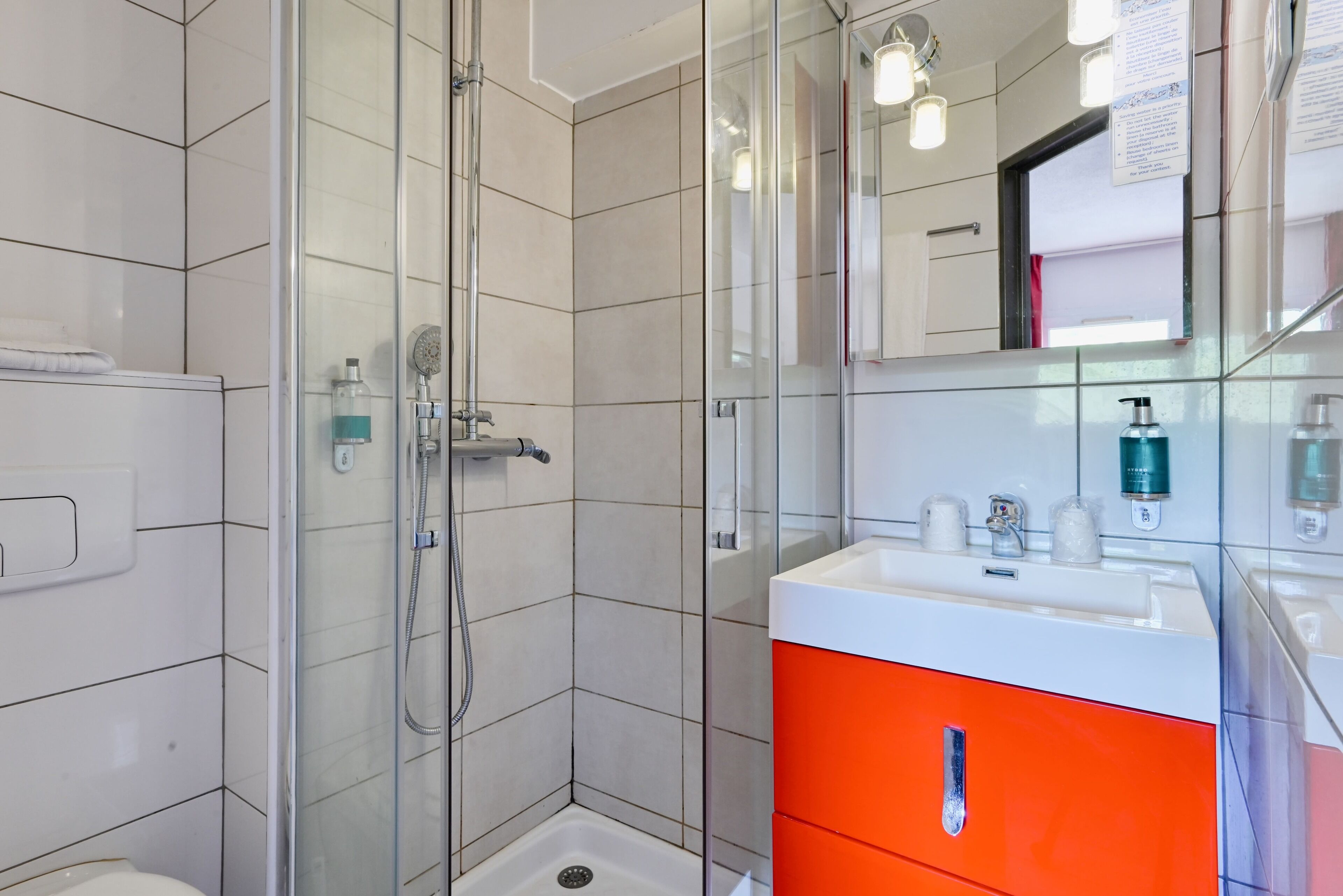 A modern bathroom with a glass-enclosed shower, white tiled walls, a white sink with orange cabinets, and a mirror above the sink.