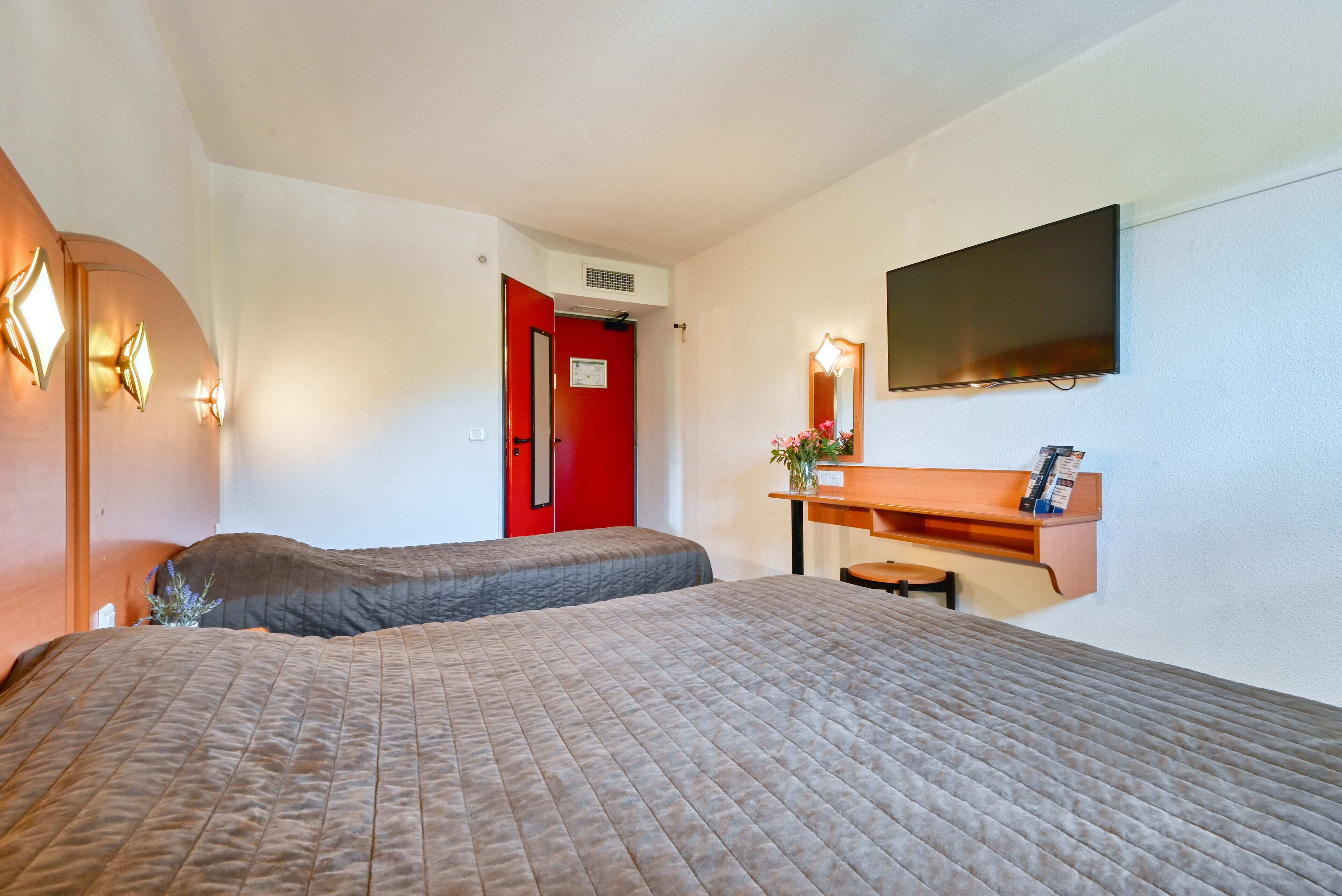 Une chambre d’hôtel avec deux lits, une porte rouge, un téléviseur à écran plat et un petit bureau agrémenté d’un miroir et d’un vase de fleurs.
