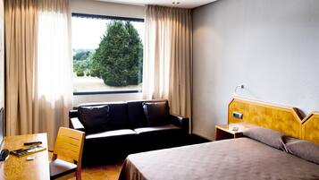 A hotel room with a bed, a sofa, a desk with a computer and remote control, and a window with curtains.