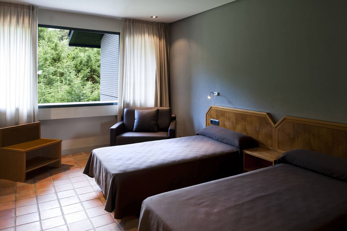 A room with two beds, a chair, a small table, and a window with curtains.