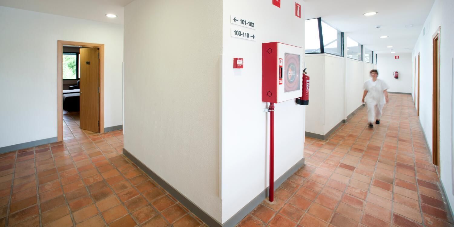 Un pasillo con suelo de baldosas, un extintor de incendios montado en la pared y señales direccionales que indican los números de las habitaciones.