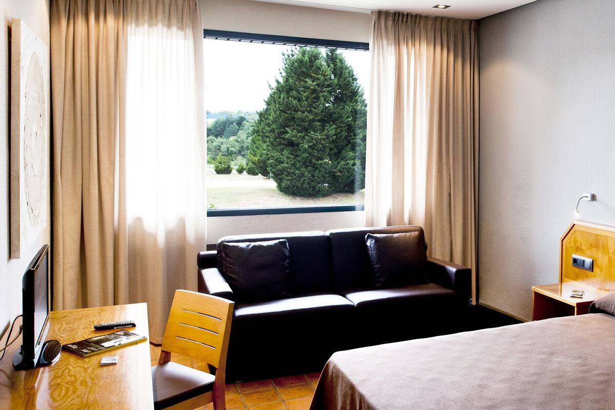 A hotel room with a bed, a sofa, a desk with a computer and remote controls, a nightstand, and a window with curtains.